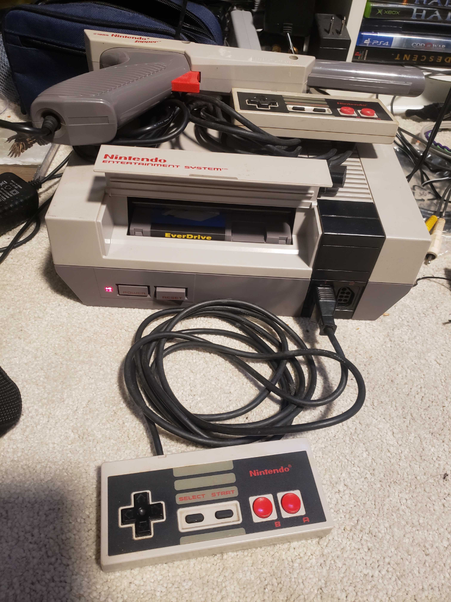 An NES Console with the flap opened up. On top is a Zapper gun with another controller, both with cables neatly wrapped up. On the floor, plugged in, in front of the NES, is another controller. The Power LED is on.